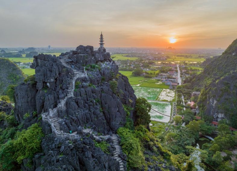 Scenic view from Hang Múa in Ninh Bình, Vietnam, showing winding stairs, lush rice paddies, rivers, and towering limestone formations.